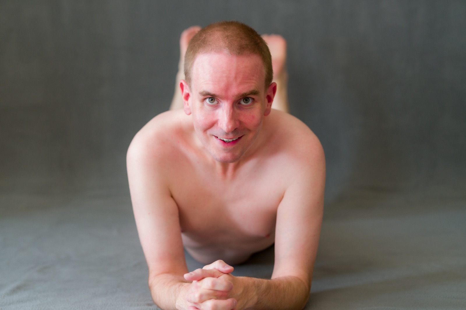 SJ Nude lying on his stomach, chin on clasped hands, gray backdrop, San Jose